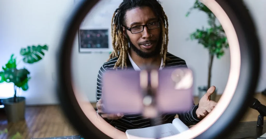 Content creator with dreadlocks and glasses speaks to a smartphone mounted in a ring light, gesturing in a bright, plant-filled room.