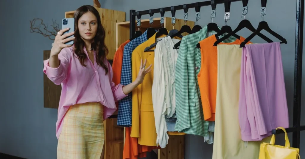 Young woman in pink blouse takes a selfie beside a clothing rack filled with colorful garments, conveying a stylish, cheerful mood.