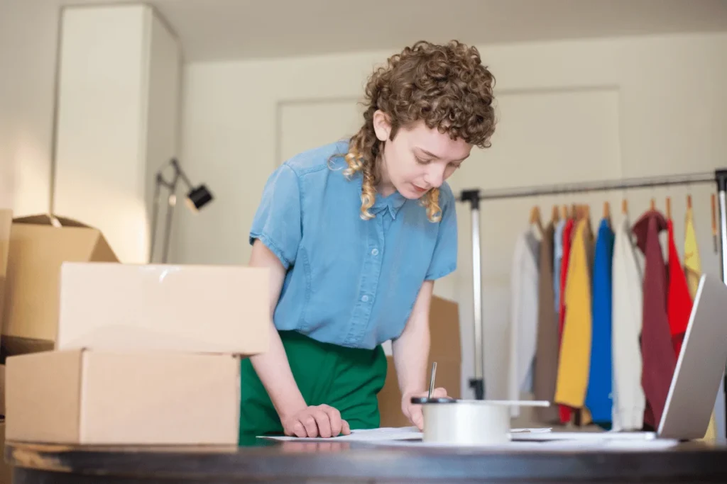 Focused young woman in a blue shirt and green pants works at a table beside shipping boxes, laptop, and a clothing rack in a bright room.