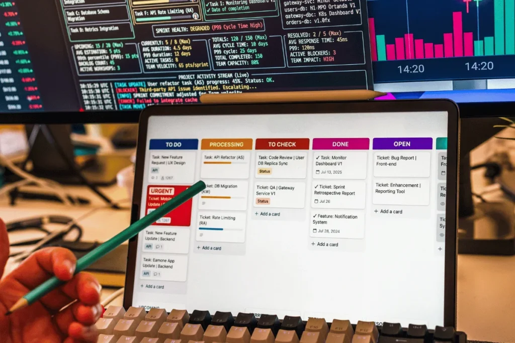 Close-up of a developer&rsquo;s desk: a tablet shows a color-coded task board, with code and charts on monitors behind it, creating a busy tech-work mood.