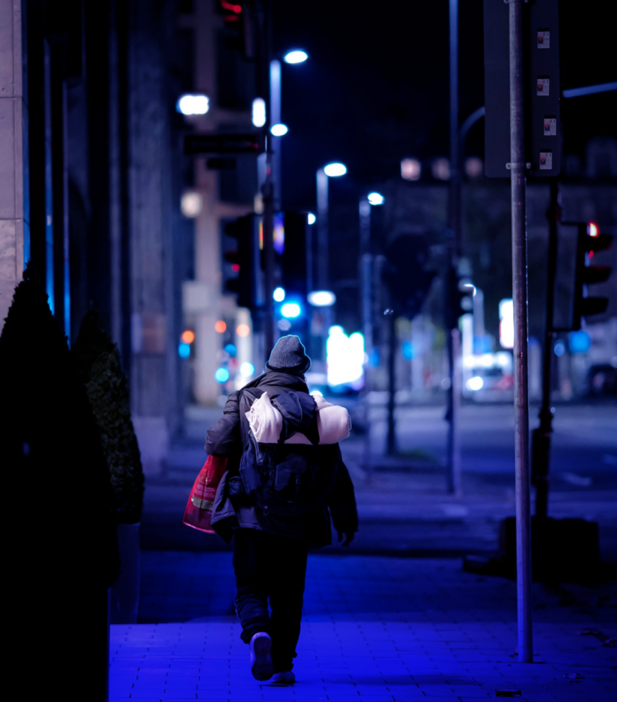 A man wearing a grey beanie and black baggy clothes is walking by night in New York streets, carrying several bags including one with a duvet in it.